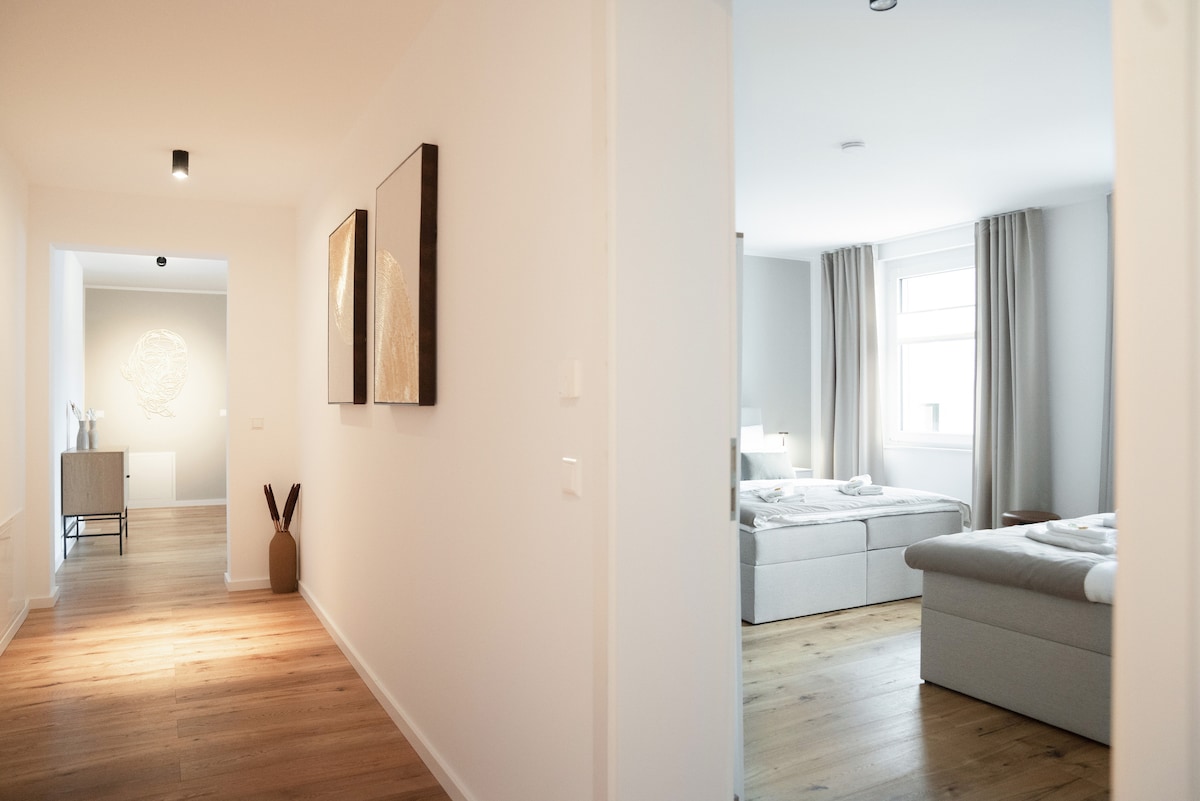 A softly lit hallway features wooden flooring leading to an adjacent room. On one side, abstract art pieces are displayed on white walls, while the other side opens to a spacious bedroom with grey curtains framing the windows and inviting natural light.