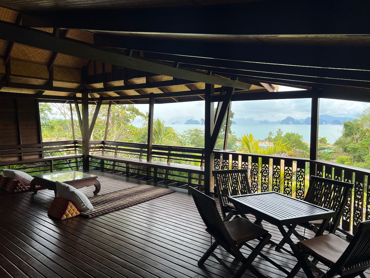 An open-air deck offers panoramic views of Phang Nga Bay, with a spacious wooden dining table and chairs arranged beneath a sloping roof. Cushioned seating provides a relaxing spot, while tropical greenery surrounds the area, creating a serene atmosphere.