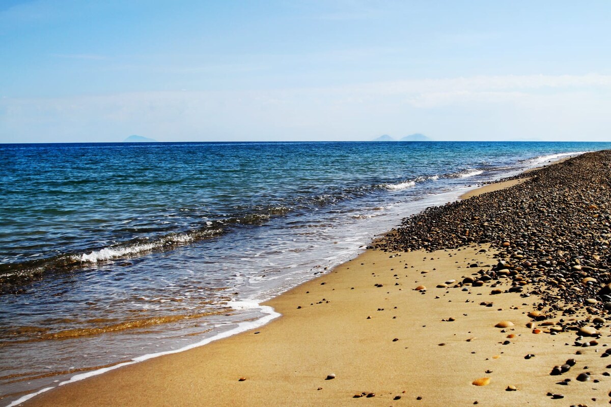 A serene beach scene is captured, featuring a soft, sandy shoreline leading to gentle waves lapping against the shore. The water transitions from deep blue to light turquoise, with small pebbles scattered along the beach. In the distance, faint outlines of islands are visible on the horizon.