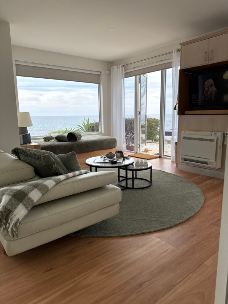 A light-filled open-plan living area features a comfortable white leather sofa adorned with a textured throw. A circular rug anchors the space, while large sliding doors provide unobstructed views of the ocean. A round coffee table holds decorative items, enhancing the serene ambiance.