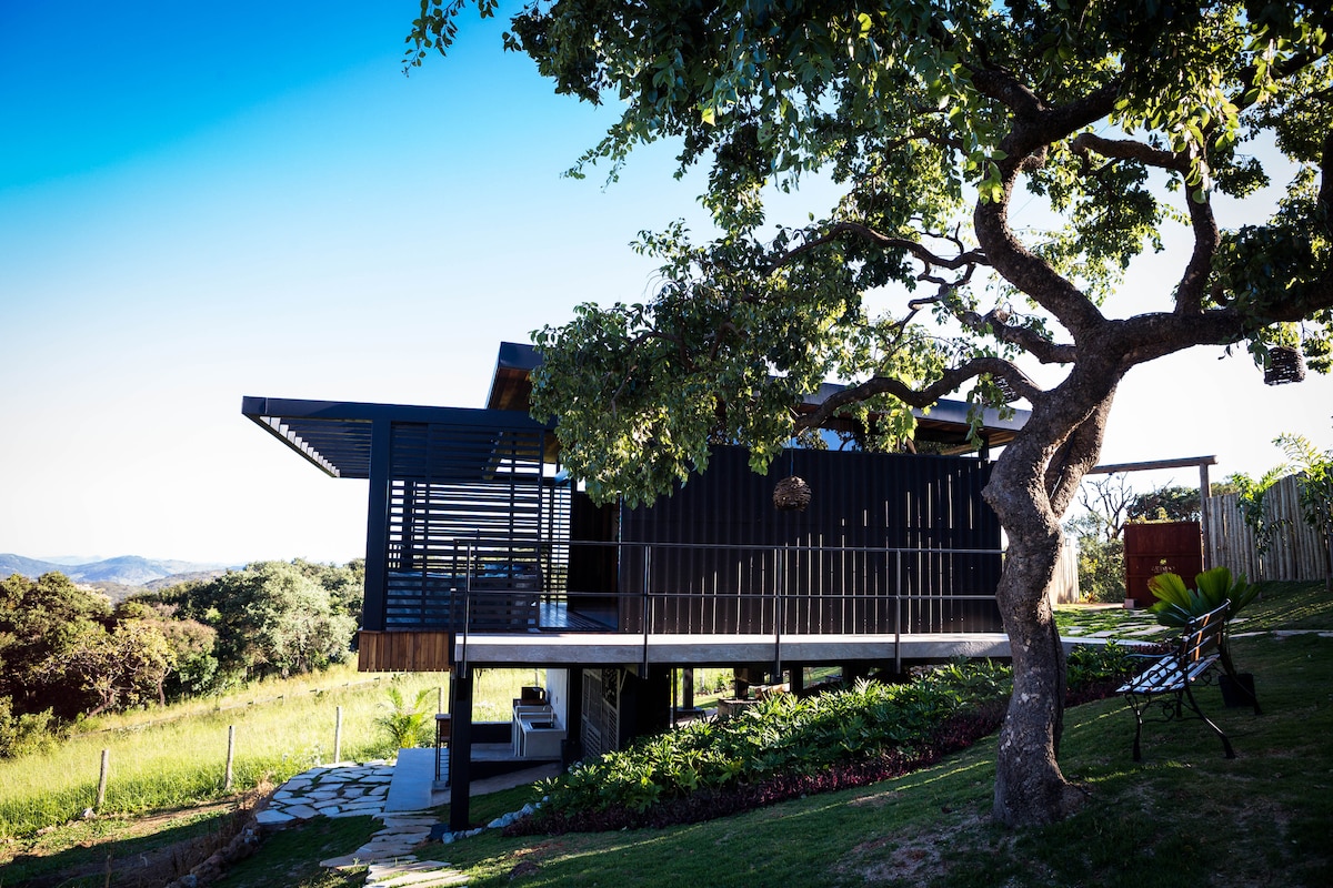 A modern cabin is nestled among greenery, showcasing a sleek design with a covered deck that extends outward. Large windows reflect natural light, while a mature tree provides shade beside the structure. The open landscape features grassy areas and distant hills.