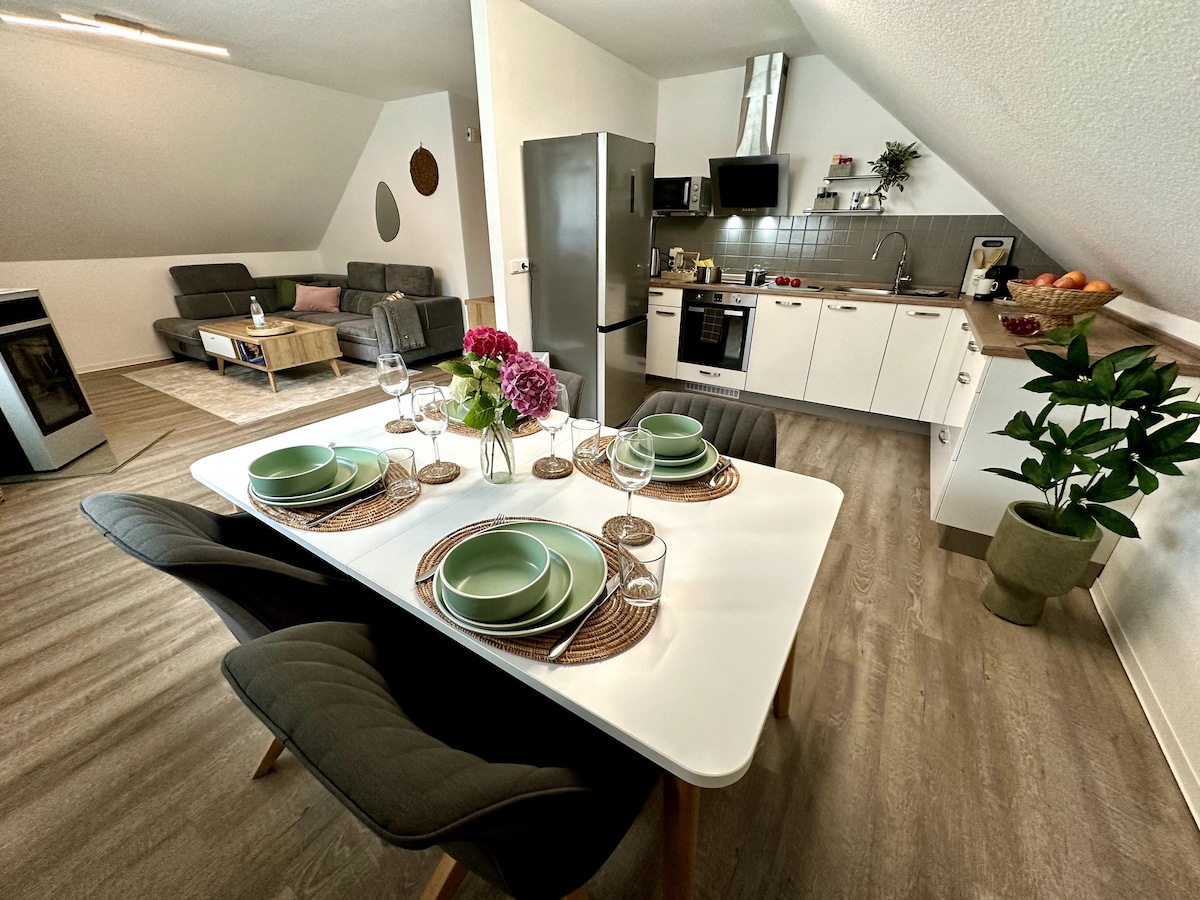 A modern kitchen and dining area is showcased, featuring a white table set for four with green dinnerware. A gray sofa and coffee table are visible in the background, along with a well-equipped kitchen displaying stainless steel appliances and a variety of fresh fruit.