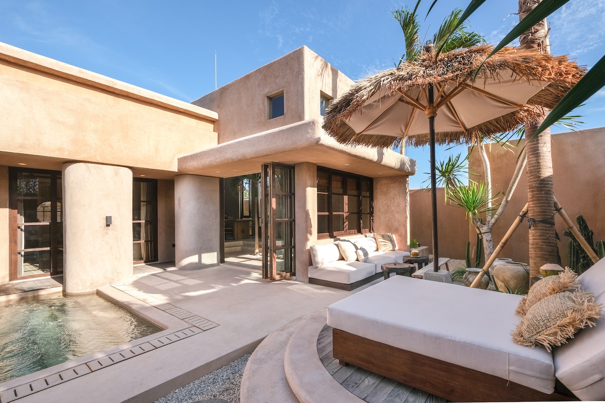 An inviting outdoor area features a private pool and a spacious sunbed nestled against adobe-style architecture. A large umbrella provides shade for lounging, while palm trees and tropical plants enhance the serene atmosphere of this open-air space.