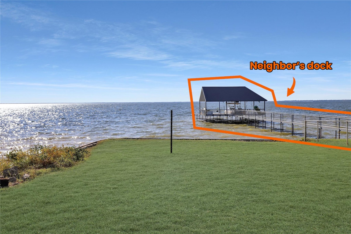 A serene view of the lake is captured, showcasing calm waters and a clear blue sky. Lush green grass leads toward the lake, with the neighbor's dock visible in the distance. The area offers a tranquil environment, ideal for outdoor relaxation and activities.