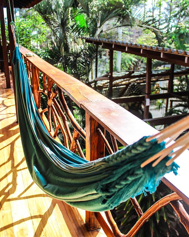 A hammock hangs peacefully from a wooden railing, surrounded by lush greenery. The natural wood of the railing and the vibrant green of the hammock create a serene setting, inviting relaxation and a connection with the surrounding garden.