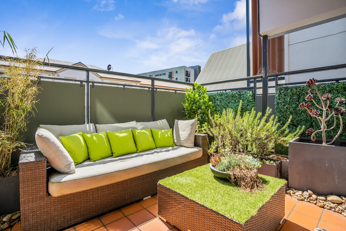 An inviting balcony area features a comfortable outdoor sofa with bright green cushions. Surrounding the seating are lush plants and a small table adorned with greenery, offering a serene space for relaxation. The backdrop showcases a clear blue sky and nearby buildings.