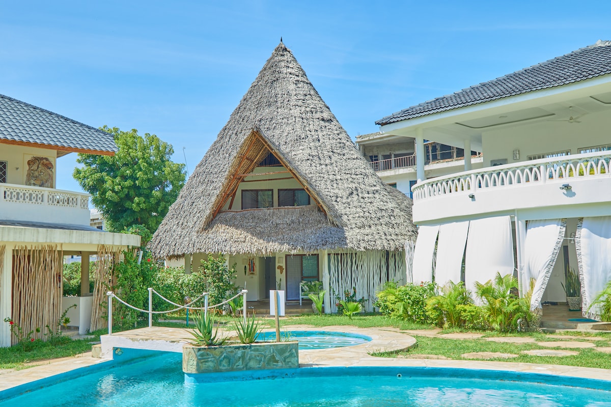 A thatched-roof beach house is surrounded by a stone pathway and lush greenery. Two sparkling swimming pools are visible, framed by tropical plants. The clear blue sky enhances the serene atmosphere of the outdoor space, inviting relaxation and enjoyment of the coastal environment.