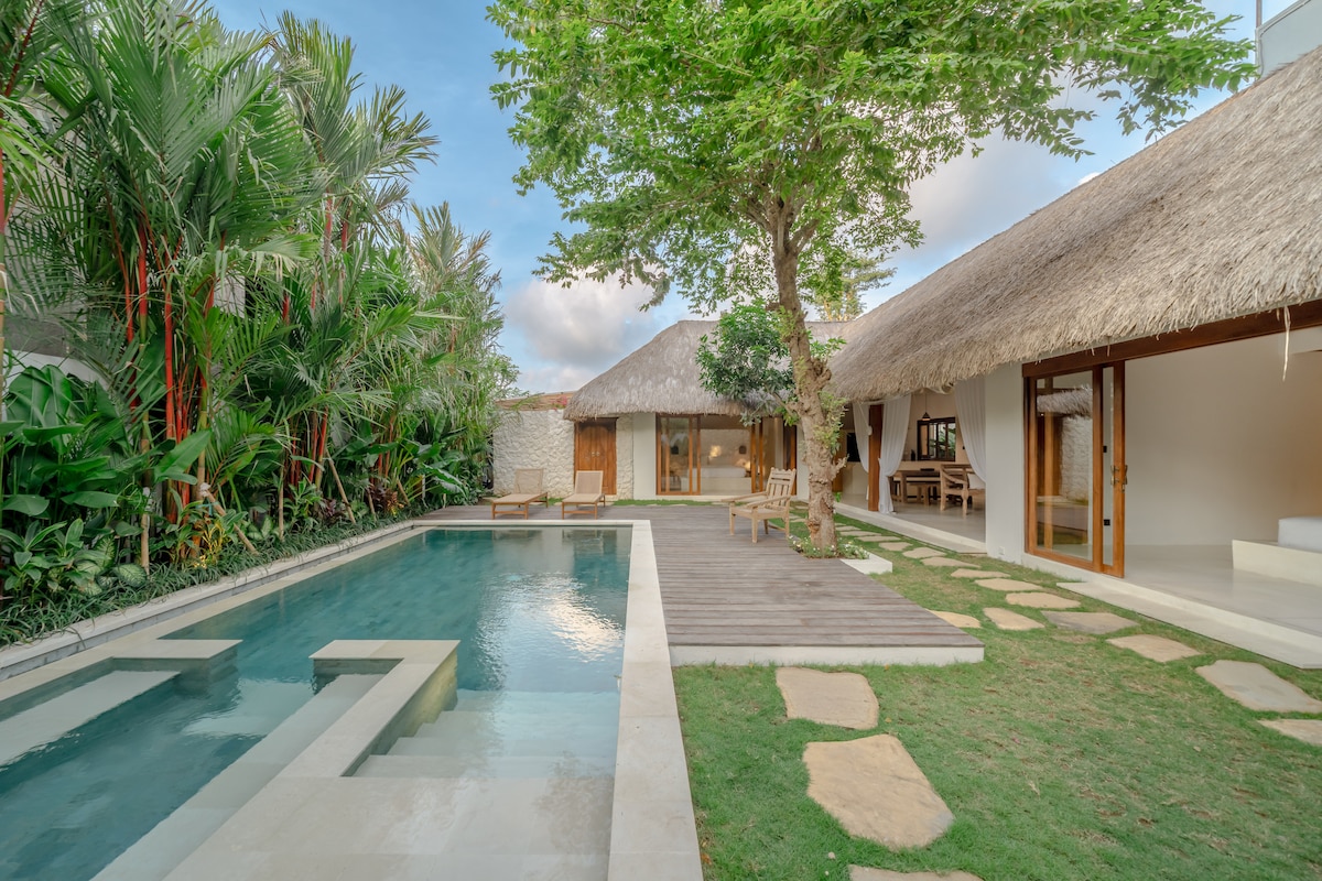 A serene outdoor area is illustrated, featuring a spacious private pool surrounded by lush tropical gardens. A wooden deck adjoins the pool, providing seating options. The design includes a traditional thatched-roof structure and large glass doors that connect indoor and outdoor spaces.