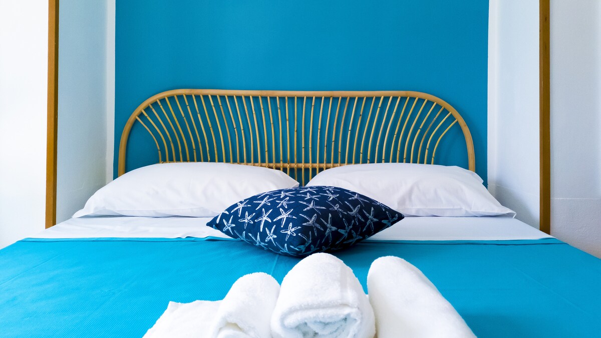 A cozy sleeping area is highlighted by a rattan headboard against a vibrant blue wall. A decorative pillow with a starfish pattern rests on a neatly made bed with white linens. Freshly folded towels are arranged at the foot of the bed.