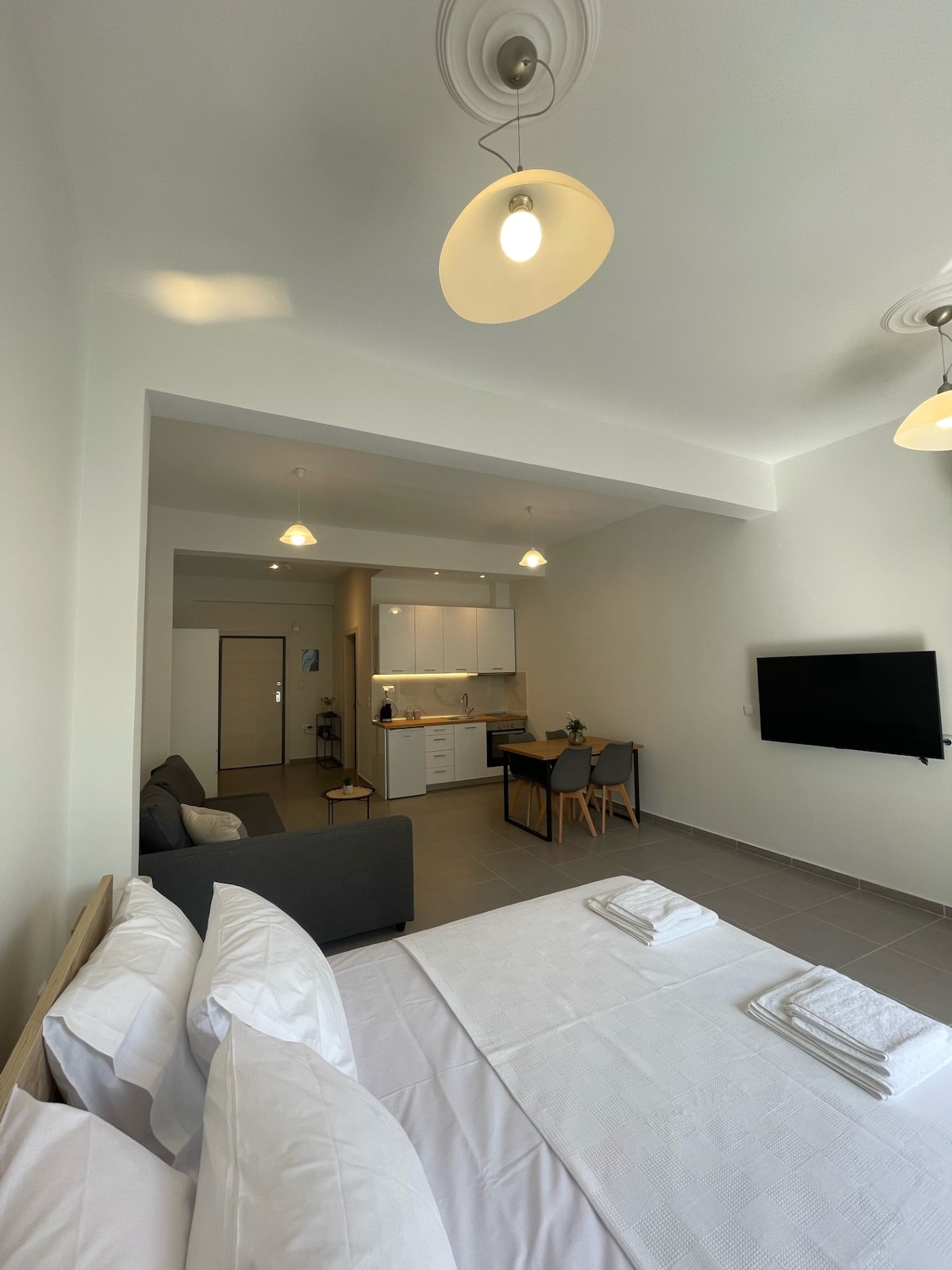 A spacious studio apartment is showcased, featuring a comfortable sleeping area with neatly arranged linens. A modern kitchenette is visible in the background, accompanied by a small dining table and chairs. Soft lighting is provided by pendant fixtures, enhancing the clean and minimalist decor.