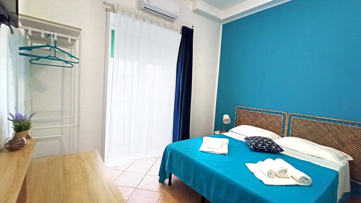 A bright bedroom features a blue accent wall, a spacious bed adorned with fresh linens and towels. A wooden dresser provides additional storage. Natural light streams in through a sheer curtain, while a small closet area with hangers is visible, offering convenience for guests.