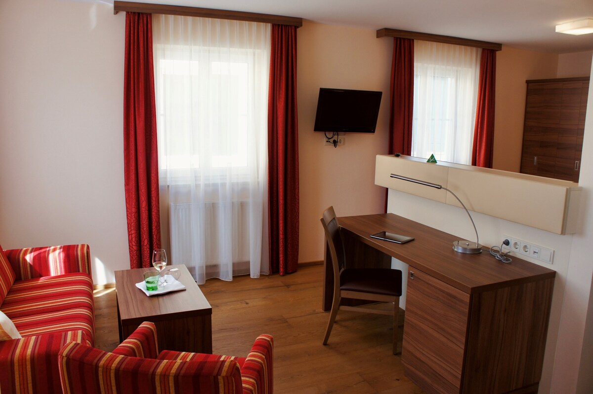 A cozy living area is furnished with a striped sofa and a wooden coffee table. A modern desk sits against the wall, accompanied by a chair. Natural light enters through floor-to-ceiling windows, accentuated by elegant curtains. A wall-mounted television is positioned for viewing.