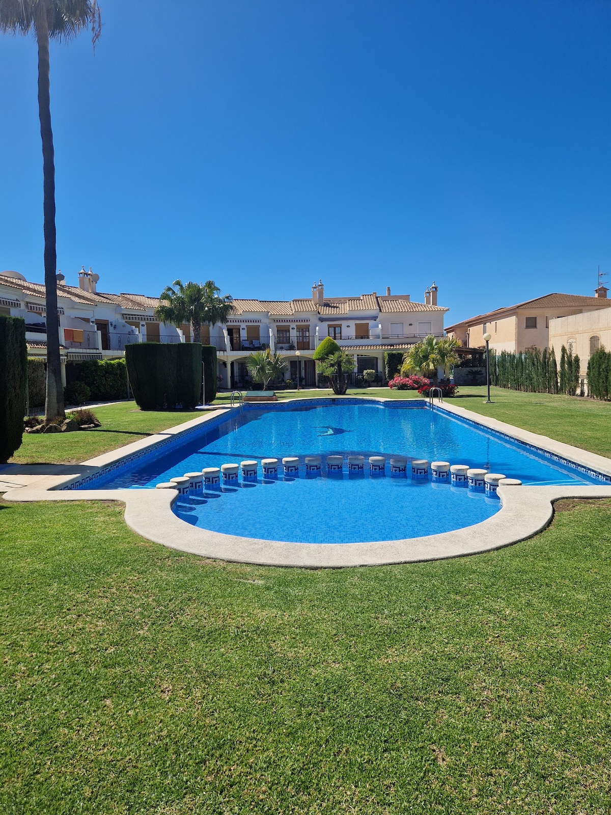 A large swimming pool is featured within a well-maintained garden area, surrounded by neatly trimmed hedges and palm trees. The pool includes a circular seating area, with clear blue water under a bright sky. The residential buildings are visible in the background.