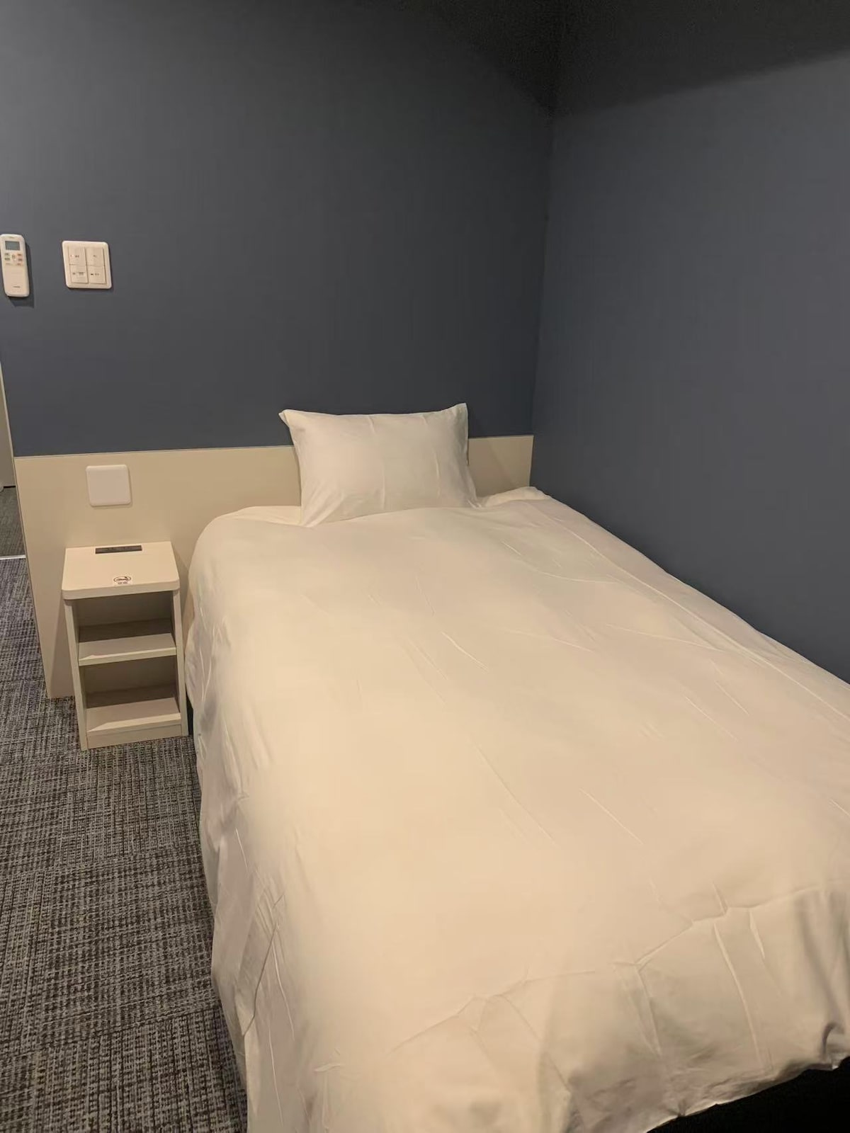 A neatly arranged sleeping area features a compact single bed dressed in crisp white linens. A bedside table is positioned next to the bed, providing convenience for personal items. The wall is painted in a calming blue, complementing the overall minimalistic design.