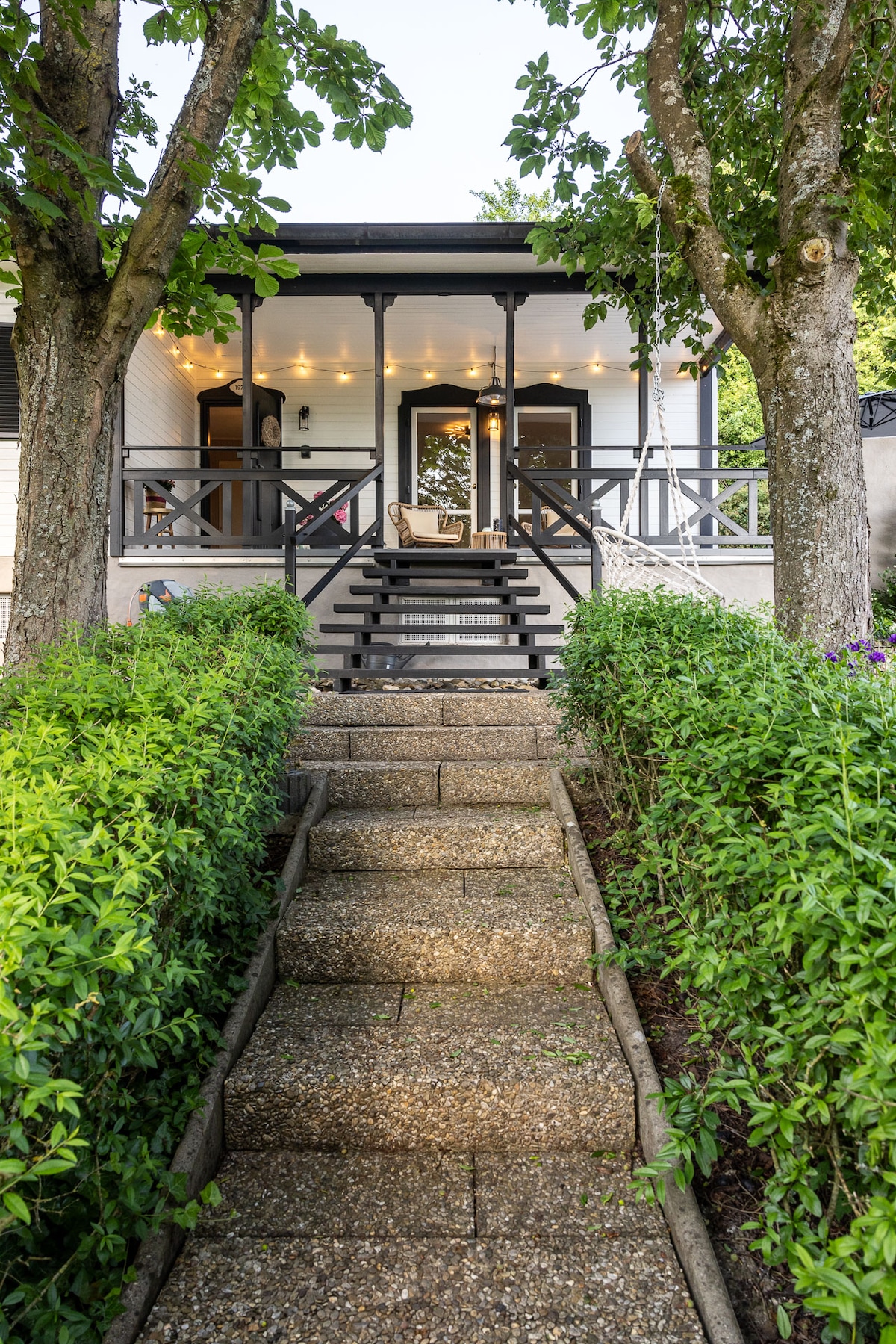 A stone path leads to the entrance of the cottage, framed by lush greenery and tall trees. The porch features a welcoming seating area adorned with string lights, creating a cozy ambiance. Stairs ascend to the entrance, inviting guests to enter and explore.