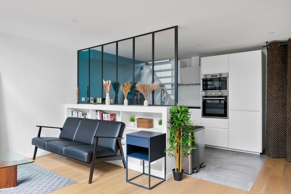 A modern living area is visible, featuring a sleek black sofa facing a glass partition that separates the living space from the kitchen. A small blue table sits beside the sofa, complemented by decorative plants and books on the shelf behind it.