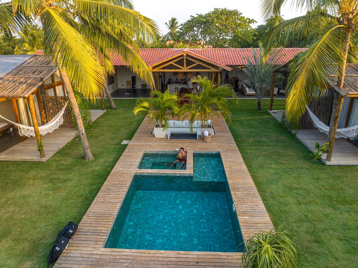 A private outdoor area features a heated jacuzzi adjacent to a swimming pool, surrounded by lush palm trees and well-maintained grass. Several thatched-roof bungalows are visible in the background, providing a serene setting for relaxation.