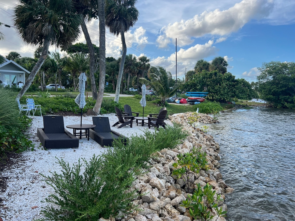 Outdoor seating is arranged on a pebbled shoreline, featuring dark wicker lounge chairs and umbrellas. Lush greenery frames the space, leading to the calm water of the Indian River, where kayaks are visible along the bank under a partly cloudy sky.