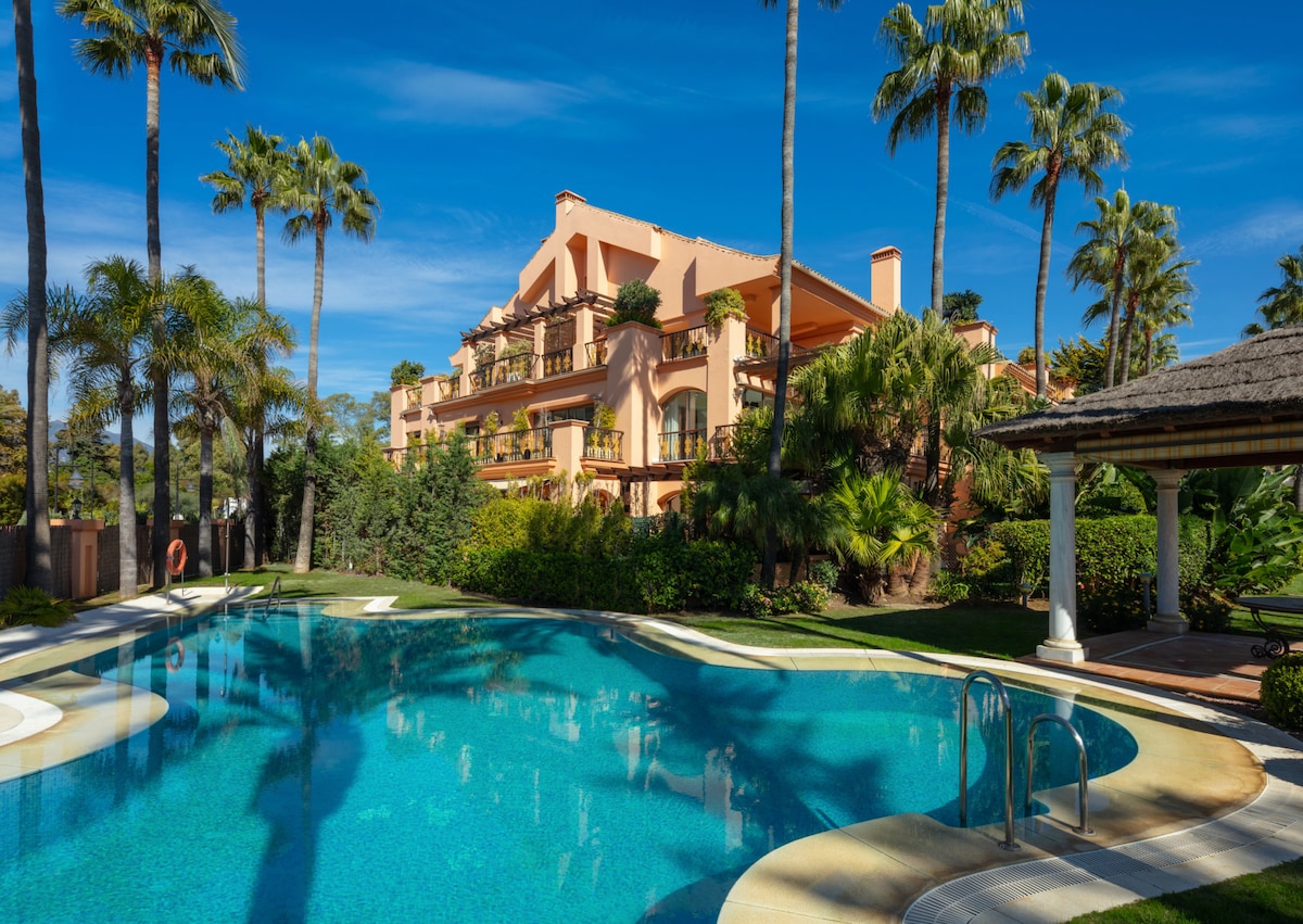 A well-maintained pool is surrounded by lush tropical landscaping and palm trees. The vibrant waters reflect the clear blue sky. The elegant apartment building can be seen in the background, featuring balconies that overlook the pool area.