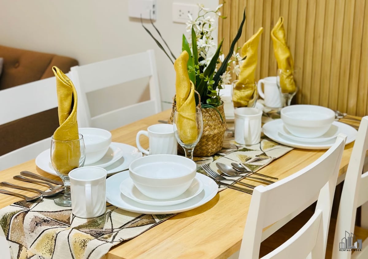 A dining table is set with elegant dishware, including white bowls, cups, and cutlery. Each place setting features golden napkins neatly arranged, and a centerpiece of greenery brings a touch of freshness to the table. The warm wooden tones of the table complement the overall design.