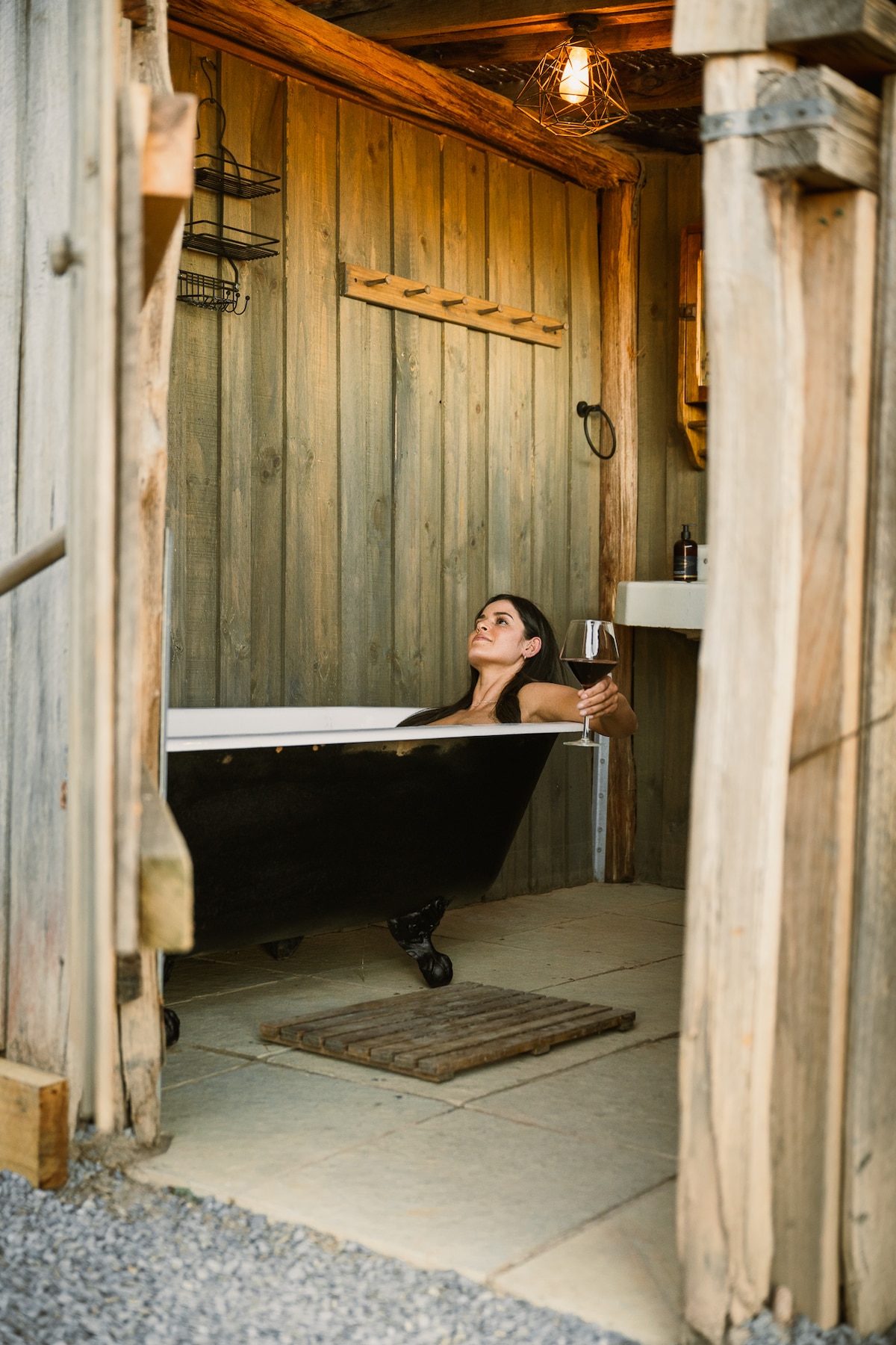 An outdoor bathroom features a freestanding black bathtub set against wooden walls, providing a rustic aesthetic. A person enjoys a glass of wine while seated in the tub, with soft lighting from a hanging bulb enriching the serene atmosphere.