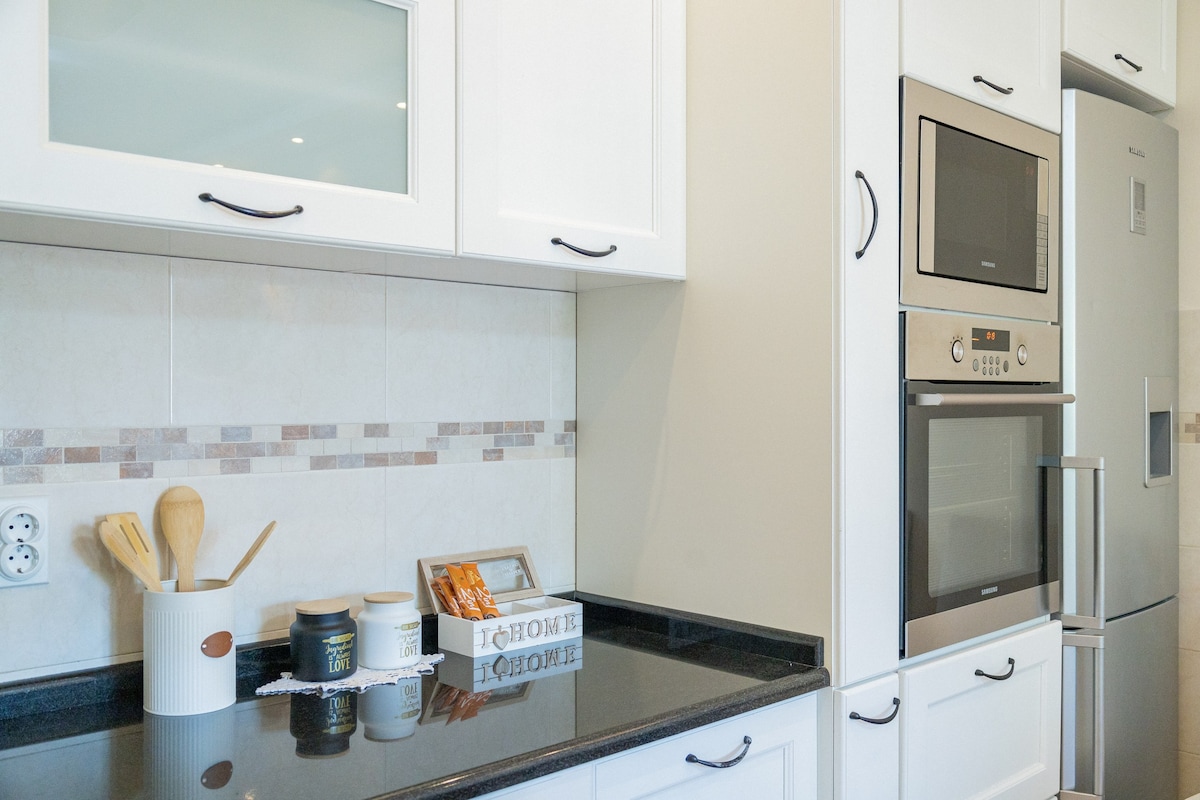 The kitchen is equipped with modern appliances, including an oven and microwave, integrated within white cabinetry. A black countertop contrasts with the cabinetry, and cooking utensils are neatly arranged in a holder. A decorative box adds a touch of organization to the space.