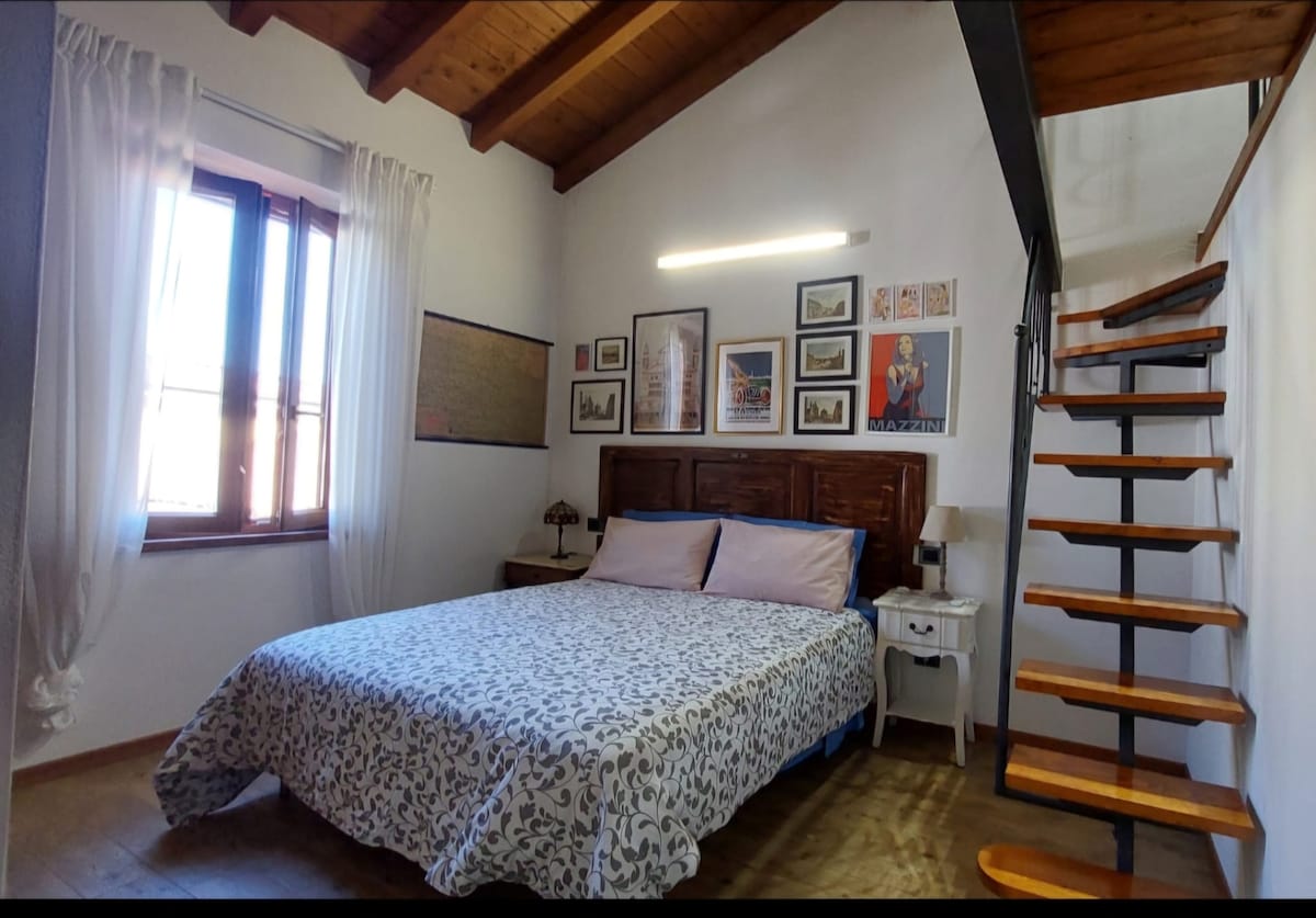 A well-lit bedroom features a spacious bed covered in a patterned blanket. The room includes a window with sheer curtains, allowing natural light to filter in. A series of framed artworks adorns the wall, while a wooden staircase leads to an upper loft area.