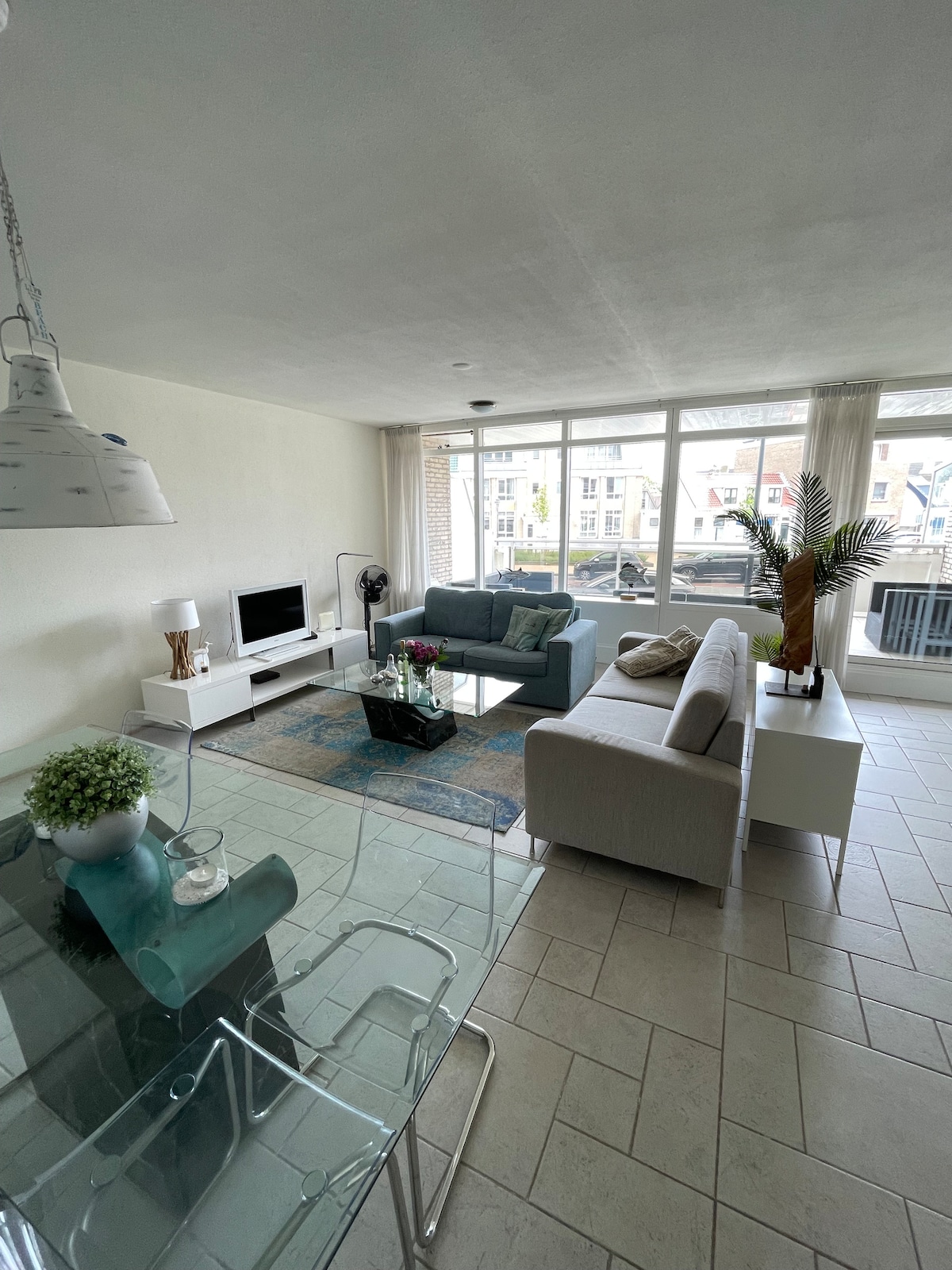 A bright living room features large windows allowing natural light to flow in. A comfortable sectional sofa is accompanied by a couple of armchairs, while a glass coffee table sits atop a textured area rug. A dining table with transparent chairs is visible, creating an open, airy feel.