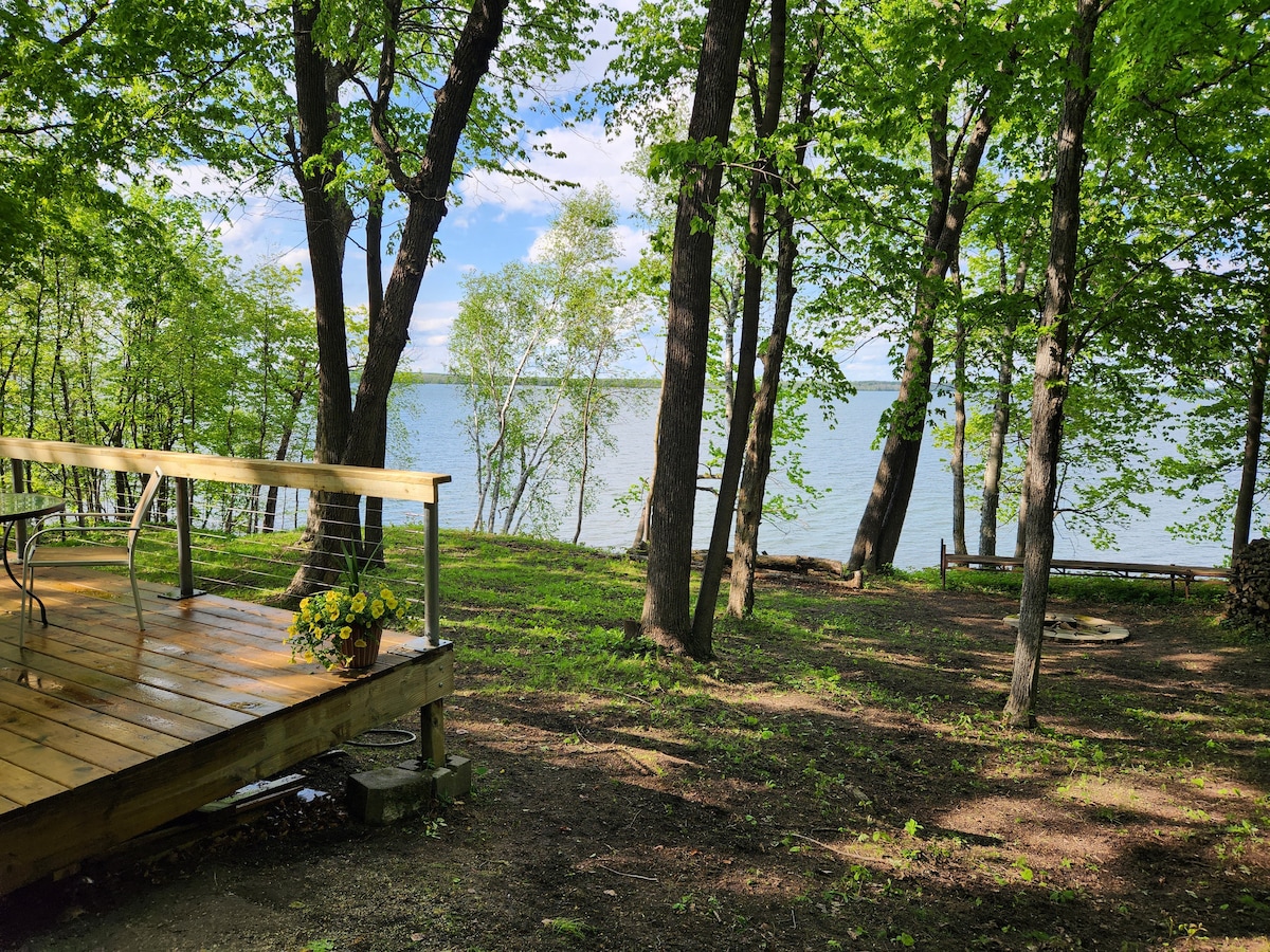 A wooden deck extends towards Shell Lake, surrounded by lush trees and greenery. The calm water is visible through the foliage, creating a serene lakeside setting. A fire pit is situated in the grassy area near the shore, enhancing the outdoor experience.