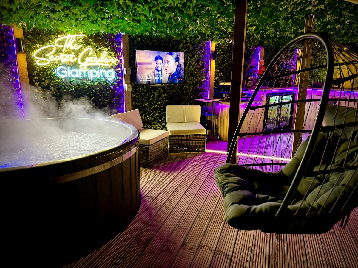 A cozy outdoor entertainment area is illuminated by colorful lights, featuring a wood-burning hot tub filled with steam. Two comfortable chairs are nestled beside the tub, and a modern swing chair enhances the inviting atmosphere. A TV is mounted on the wall, providing entertainment options.