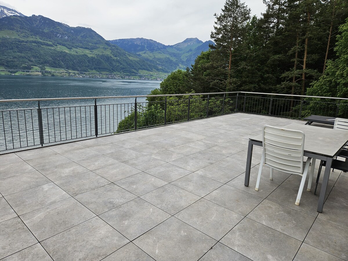 A spacious terrace features a large grey tiled surface, bordered by a railing overlooking the lake and mountains. A dining table with chairs is positioned on one side, providing a comfortable space for outdoor gatherings with scenic views.