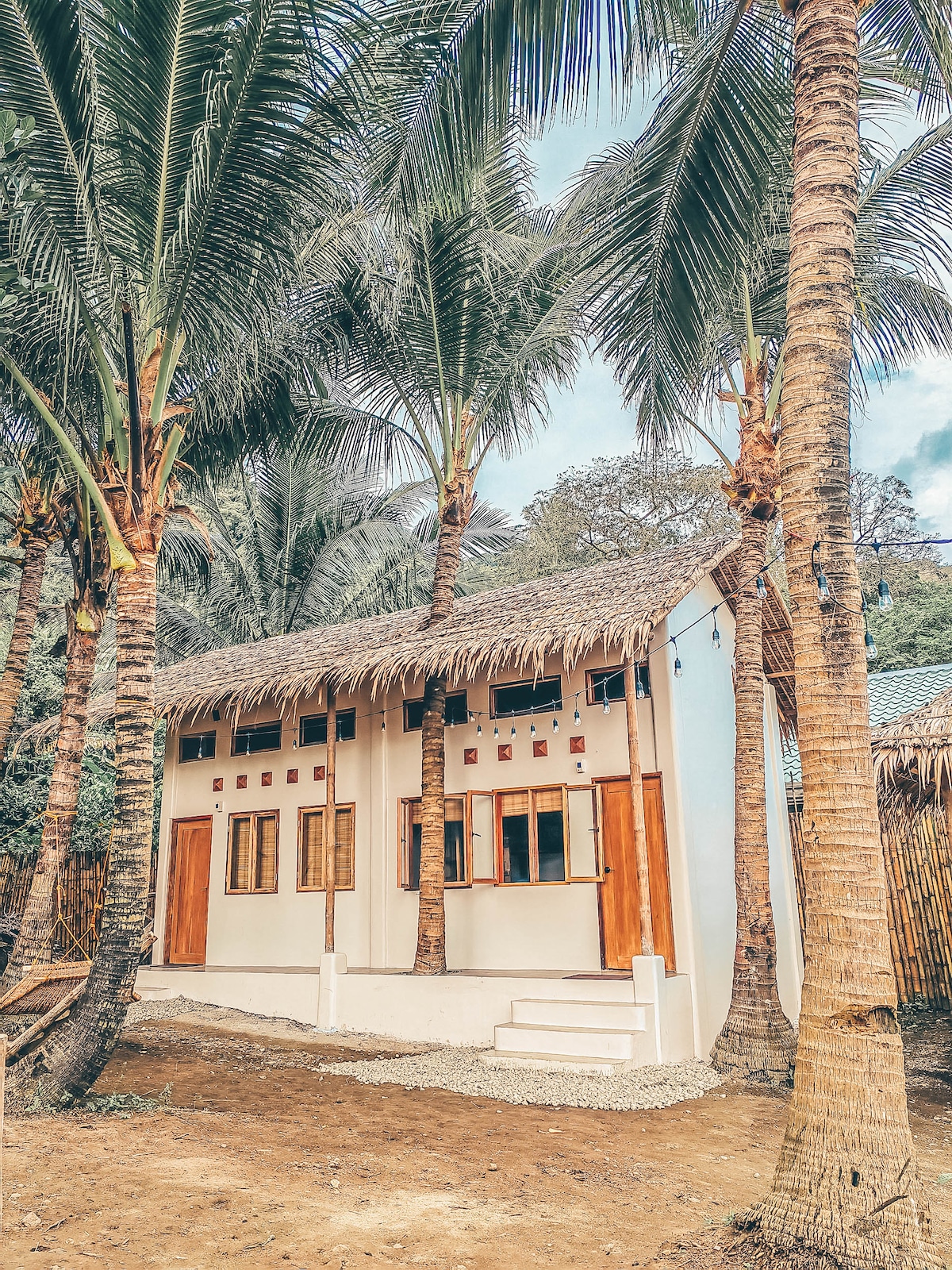 The charming cabin is surrounded by tall palm trees, featuring a thatched roof and a blend of wooden and white exterior walls. Ground-level steps lead to an inviting entryway, while several windows allow natural light to brighten the interior.