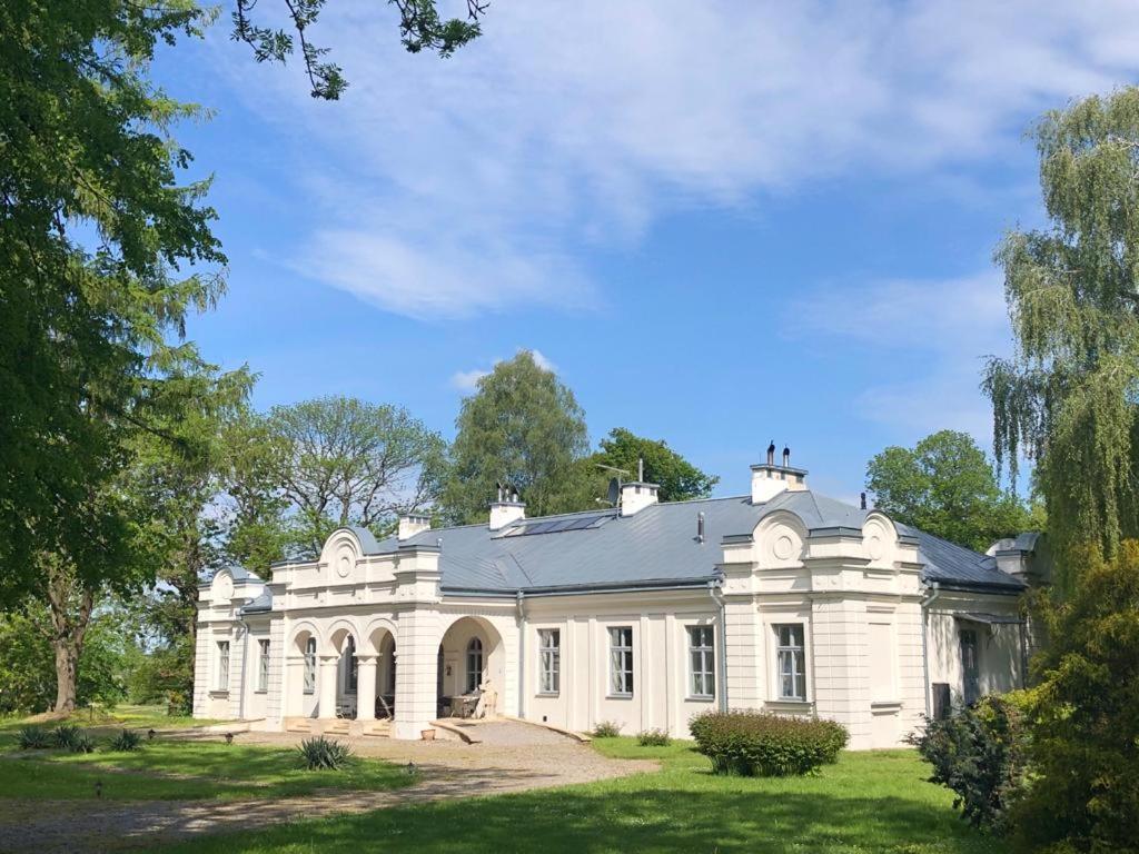 The exterior of a classic building showcases elegant architectural features, including arched doorways and symmetrical windows. Surrounded by green foliage, the structure is set against a clear blue sky, offering a serene and tranquil atmosphere in a rural setting.