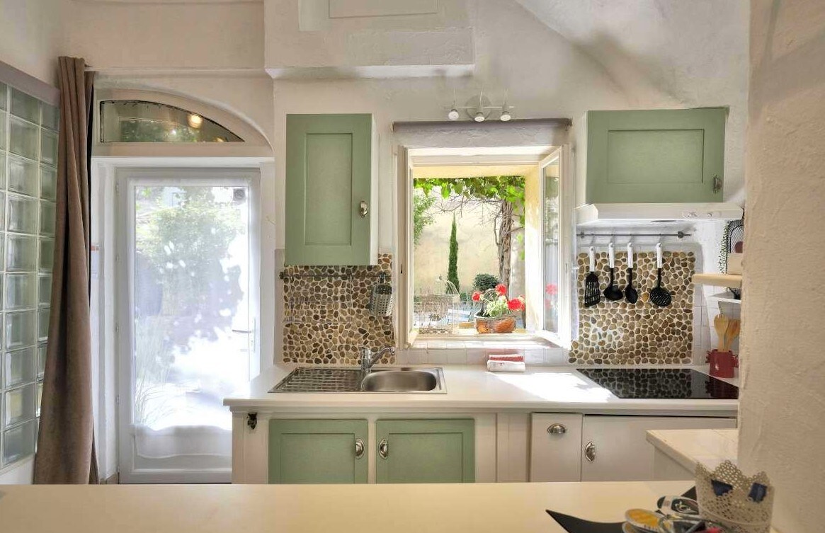 The kitchen features green cabinetry, a double sink, and a window providing natural light. A mosaic tile backsplash adds visual interest, while kitchen utensils are neatly arranged. Adjacent to the window, an outdoor view includes greenery and colorful flowers, enhancing the inviting atmosphere.