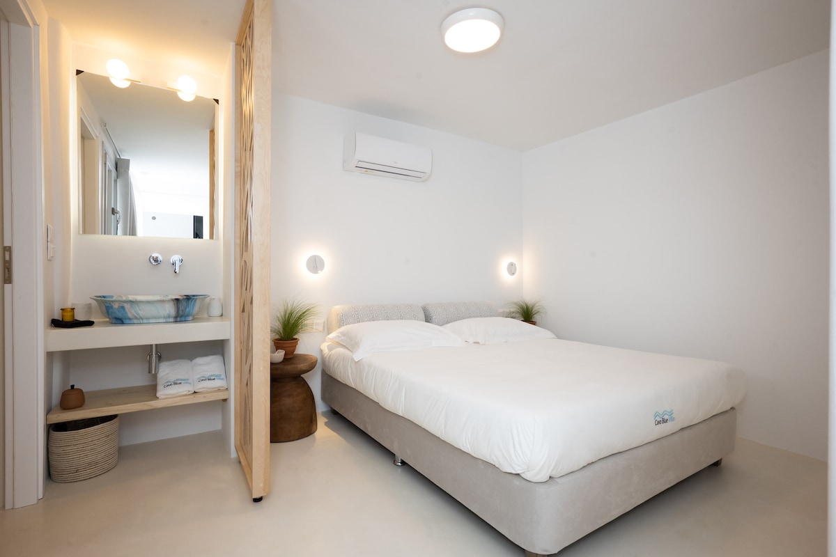 A spacious bedroom is presented with a large bed, framed by light-colored walls. A small wooden partition separates the area from a bathroom vanity, which features a blue basin and mirror. Soft illumination enhances the serene atmosphere, while decorative plants add subtle touches.