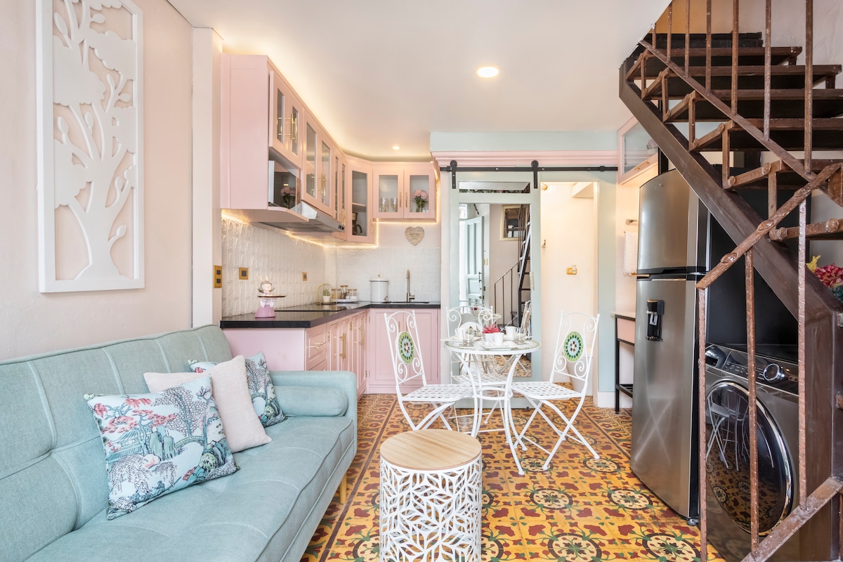 A cozy living area is complemented by a pastel-colored kitchen featuring modern appliances. A light blue sofa is adorned with decorative pillows, while a small dining table with four chairs occupies a corner. Colorful tiled flooring adds charm to the inviting space.