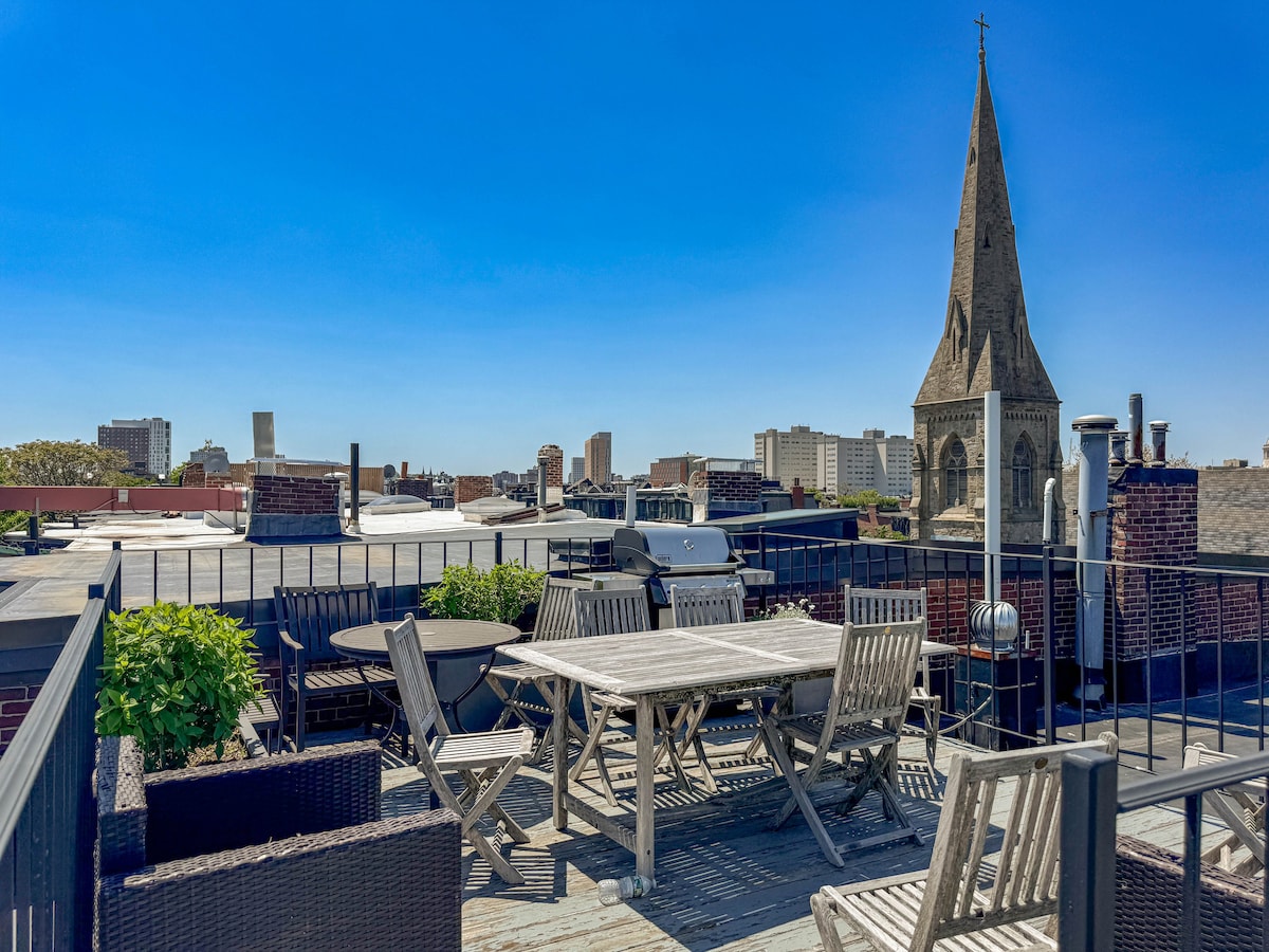 A shared roof deck is equipped with various seating arrangements, including a wooden dining table and lounge chairs. A grill is positioned nearby, with a view of the city skyline and a notable church steeple in the distance under a clear blue sky.