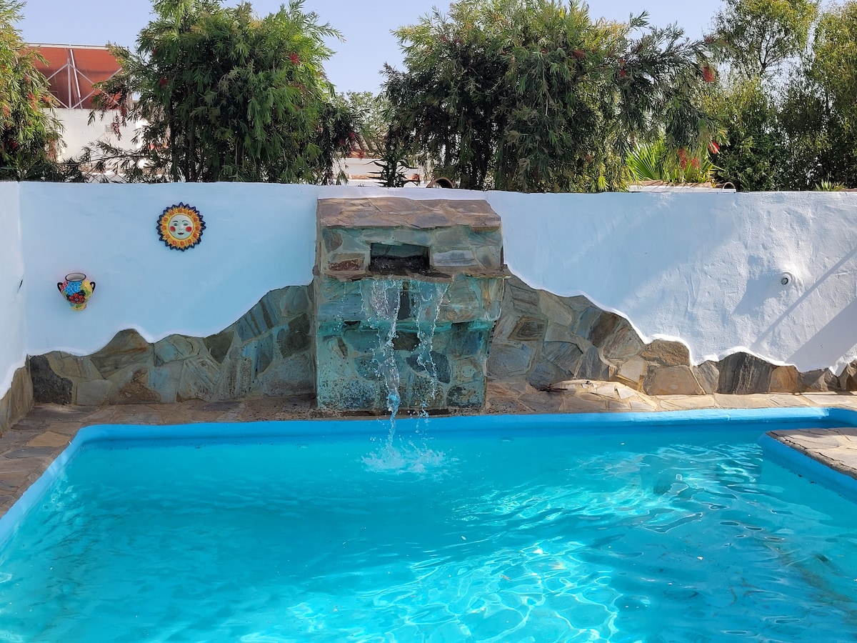 A clear blue pool features a natural stone fountain that gently cascades water. Surrounding greenery offers privacy, and decorative wall elements enhance the inviting outdoor environment. The pool area is designed for shared use, creating a serene atmosphere for relaxation.