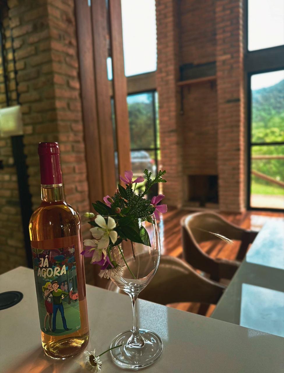 A bottle of wine is placed on a counter next to a glass, which holds a small bouquet of flowers. Natural light filters through large windows, highlighting the warm wood tones of the space and creating a welcoming atmosphere.