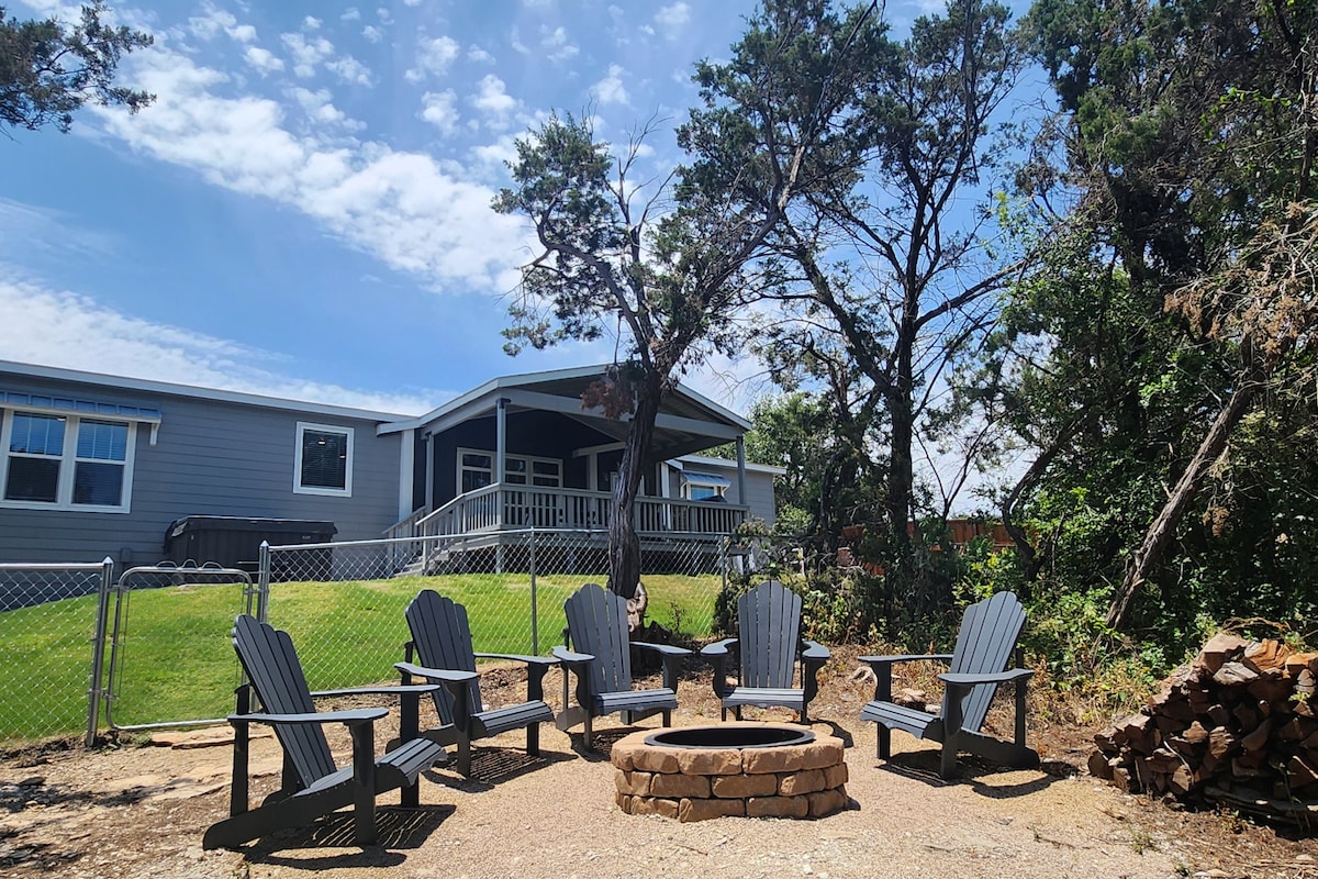 A fire pit area is surrounded by five gray Adirondack chairs, set on gravel. In the background, a home can be seen with a covered porch and large windows, while trees provide shade around the fenced yard.