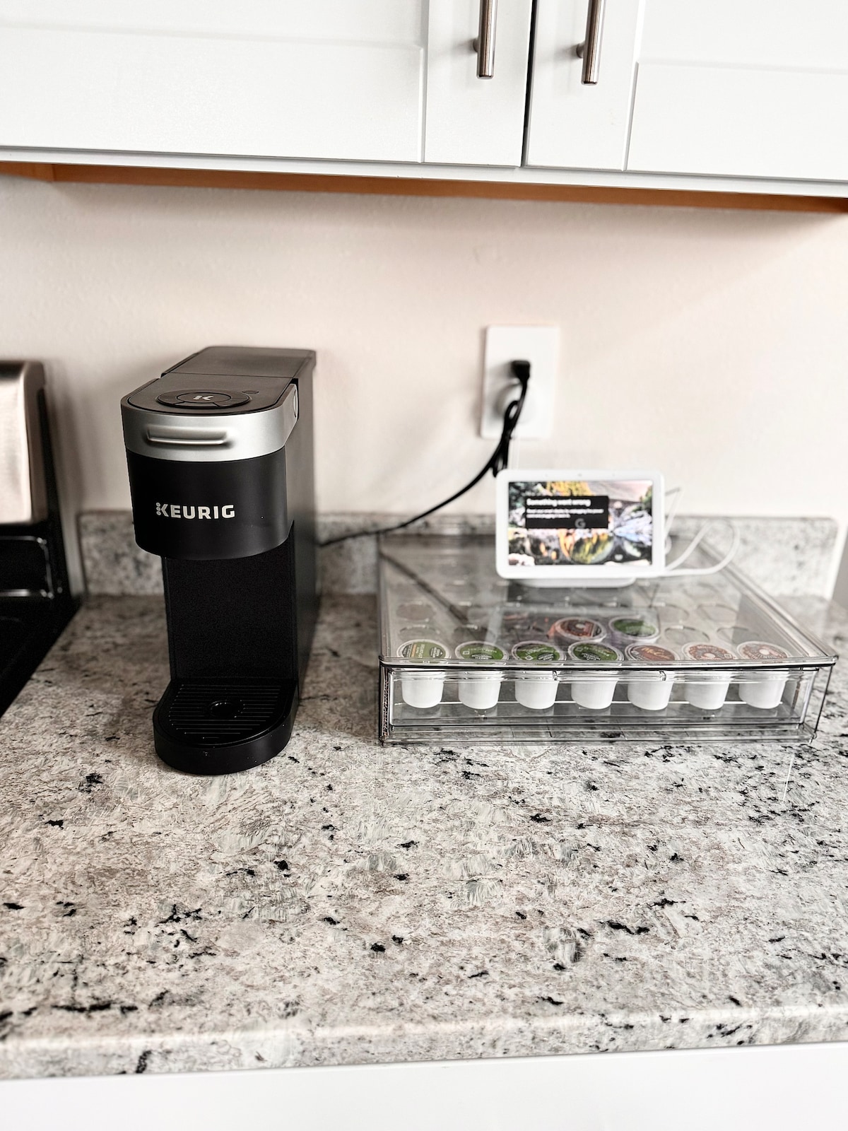 A coffee station is presented with a Keurig machine on a granite countertop. A clear container of coffee pods is organized beside it. A smartphone rests on a stand, displaying an image, while neutral cabinetry complements the setup.