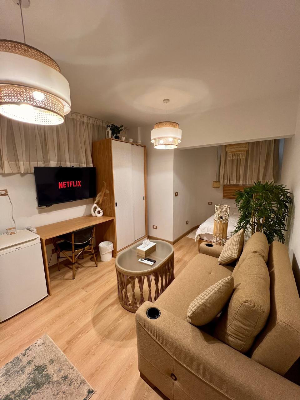 A cozy living area is depicted featuring a plush sofa with built-in cup holders. A television mounted on the wall displays the Netflix logo. A small desk with a chair is positioned near a wooden storage cabinet, while a potted plant adds a touch of greenery.