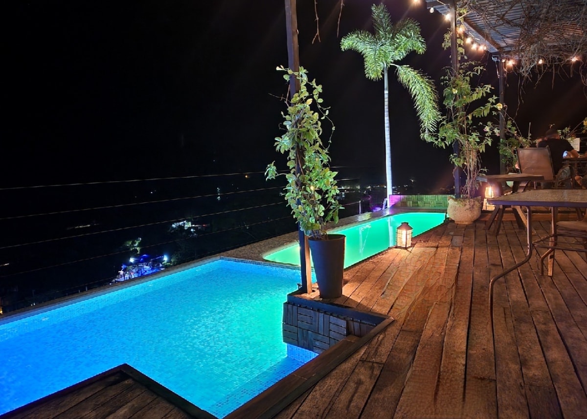 A rooftop terrace features two illuminated pools, surrounded by potted plants. Soft lighting enhances the serene atmosphere, while a wooden deck extends alongside the pools, providing space for seating. In the background, a panoramic view of the area is visible at night.