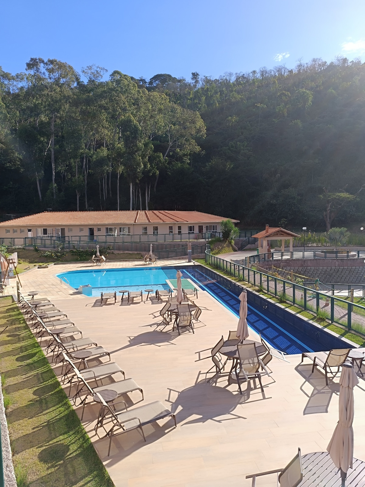 A swimming pool is surrounded by lounge chairs and umbrellas, set against a backdrop of lush greenery. A building adjacent to the pool features a warm-toned façade and a red-tiled roof. The area offers ample space for relaxation and leisure.