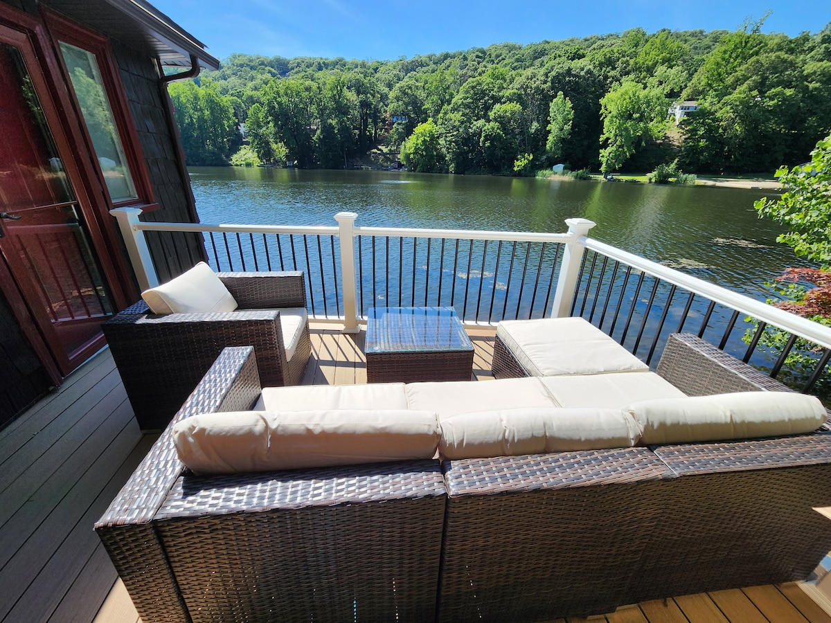 A comfortable seating area is positioned on an outdoor deck, offering a view of the calm lake surrounded by lush greenery. The furniture includes a sectional sofa and a glass-top coffee table, creating an inviting space for relaxation by the water.