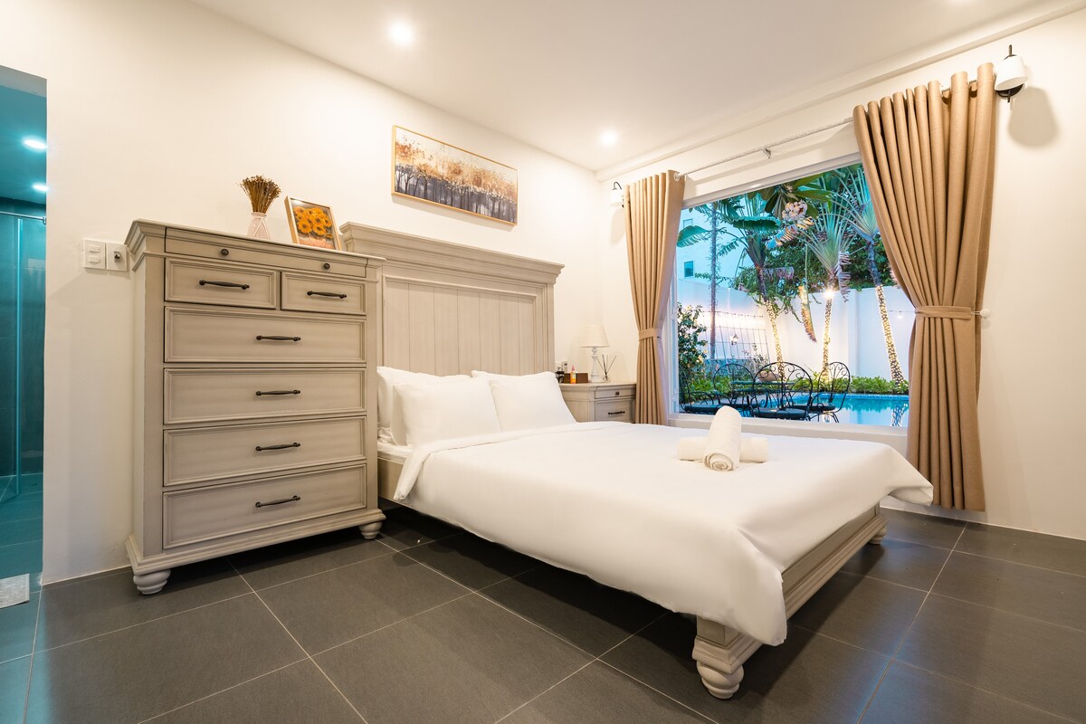 A well-appointed bedroom features a spacious bed with fresh linens and a set of towels displayed neatly. Natural light filters in through large windows, revealing a view of the garden outside. A stylish dresser complements the serene interior design.