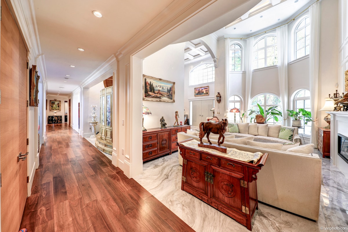 An expansive entryway leads to a bright living area featuring large windows that provide natural light. Elegant furnishings are visible, including a large sectional sofa and decorative accents. The polished wooden flooring contrasts with the marble surfaces, adding a sophisticated touch to the ambiance.
