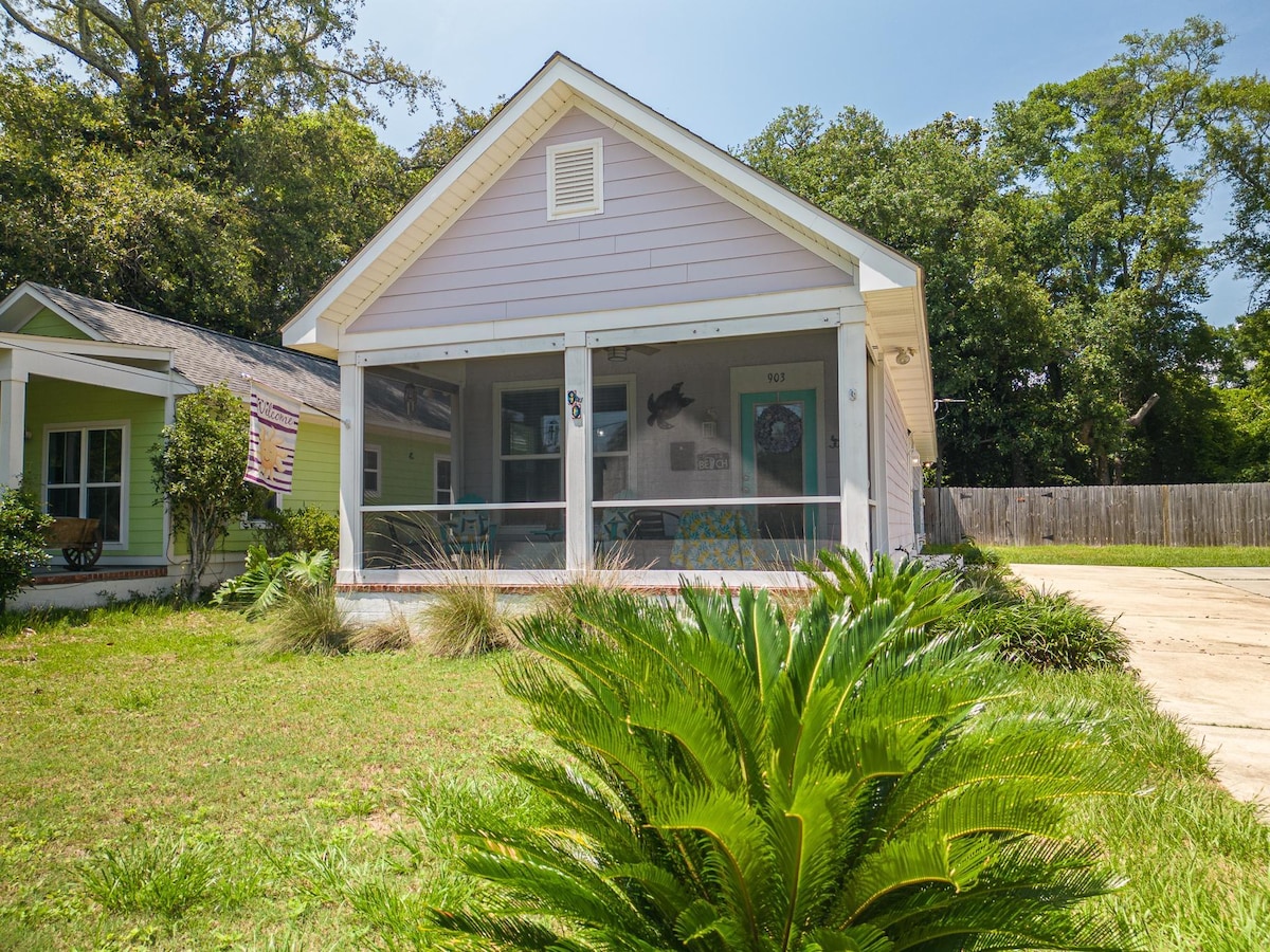 The exterior of the property showcases a charming façade in light purple, complemented by a screened-in porch. Lush greenery surrounds the home, including a large palm plant in the foreground. A spacious driveway is visible, along with neighboring houses in a serene setting.