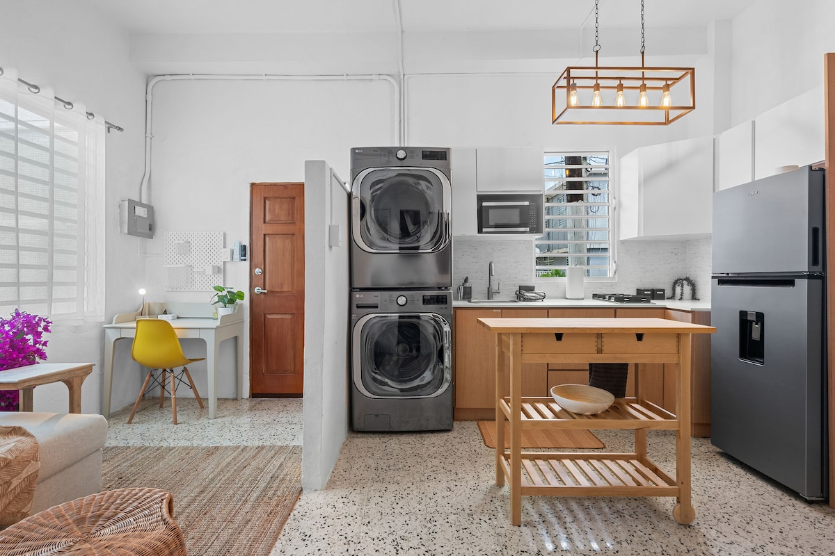 An open-concept kitchen features modern appliances, including a refrigerator and stacked washer-dryer. A wooden island with shelves provides additional workspace. A dedicated desk sits beside a window, while a small plant adds a touch of color to the light-filled area.