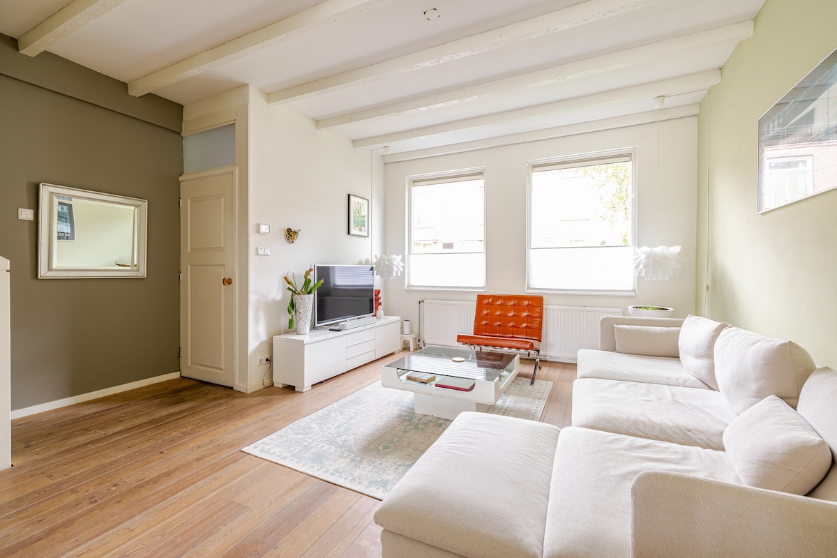 The living room features light wood flooring and a combination of white and green walls. A spacious sectional sofa is positioned alongside a modern coffee table. Large windows provide ample natural light, while a sleek television is mounted on the wall opposite the seating area.